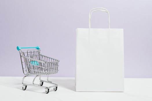 Minimalist image of a white shopping bag and mini cart on a soft pastel background.